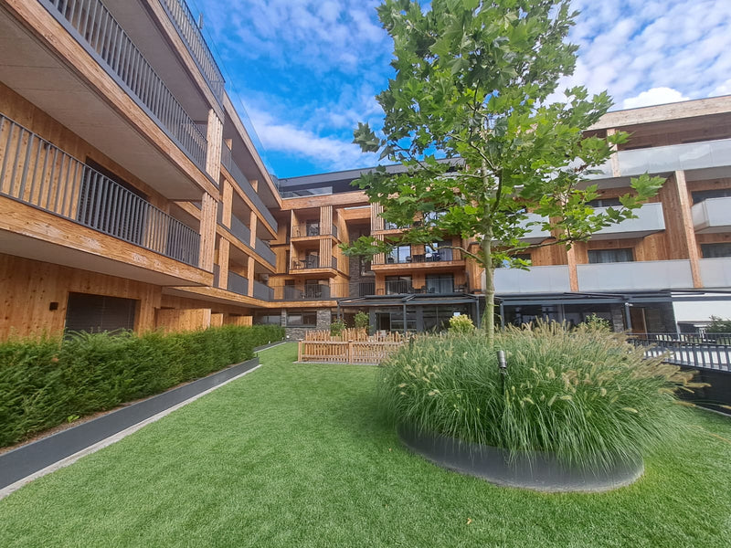 Moderner Innenhof eines Holzbau-Komplexes mit perfekter Rasenfläche und einem Solitärbaum umgeben von Ziergräsern in einem Rundbeet. Realisiert von Gartenbau Schranzhofer Kitzbühel.