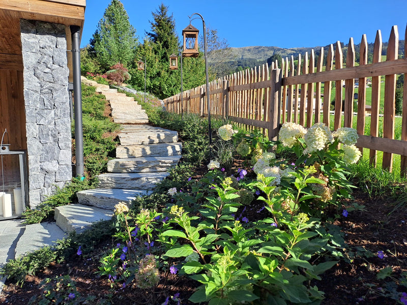 Geschwungene Natursteintreppe als Gartenweg an einem Hang, gesäumt von einem Holzzaun, Laternen und blühenden Hortensien. Wegebau und Steinarbeiten von Gartenbau Schranzhofer in Kitzbühel.