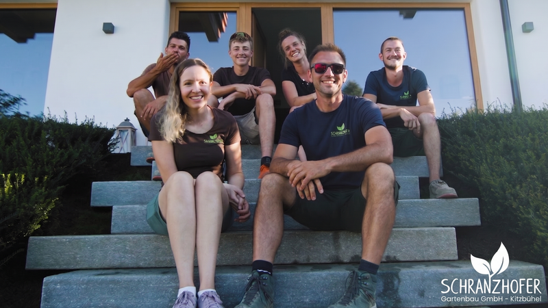 Das Team von Gartenbau Schranzhofer sitzt lächelnd auf einer Steintreppe vor dem Firmengebäude in Kitzbühel. Sympathische Landschaftsgärtner und Gartenprofis aus Tirol.
